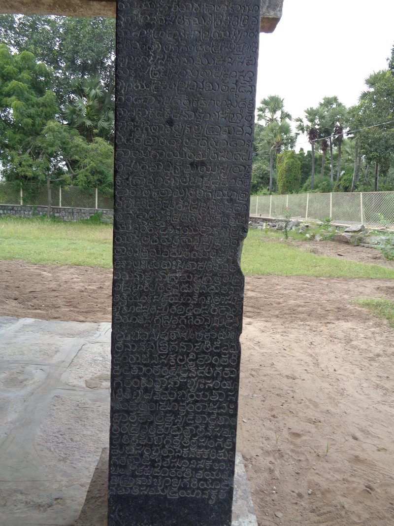 A dark granite pillar under a stone beam is covered from top to bottom with rows of engraved Telugu letters.