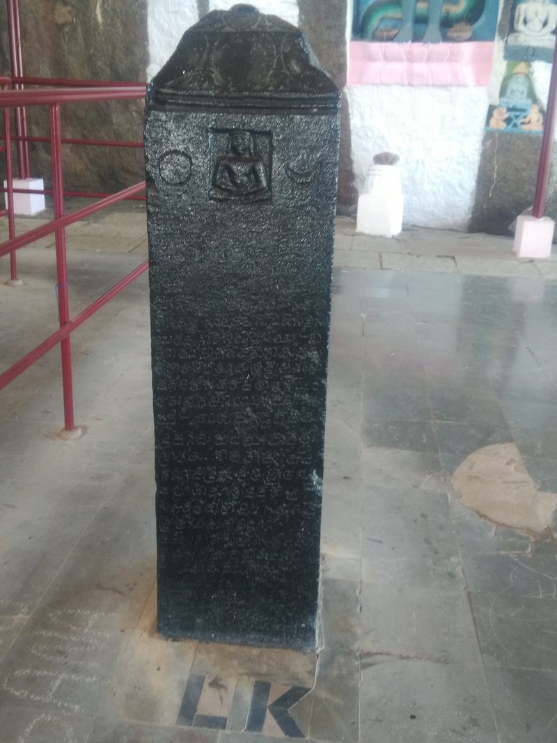 A square black pillar shows a small seated Jaina teacher in a sunken panel, with a sun disc on the left and a crescent moon on the right. Rows of letters fill the shaft below.