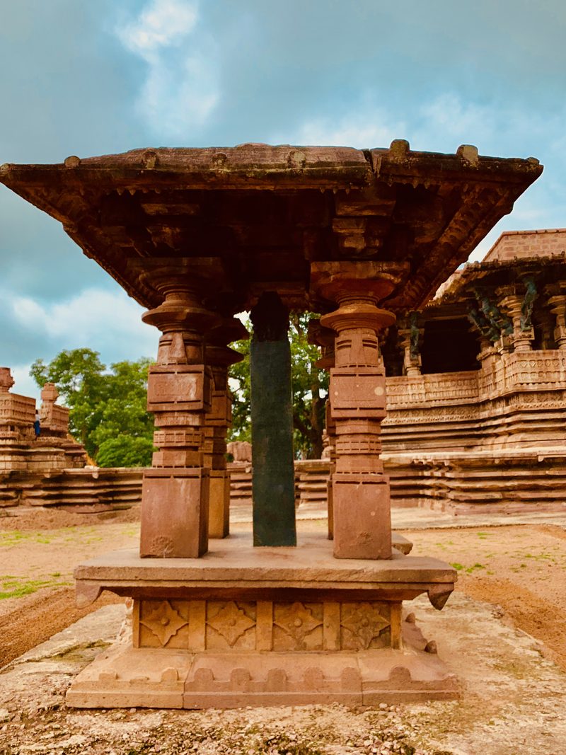 A tall black stone pillar stands inside a small open pavilion of red sandstone, with the Ramappa temple behind it.