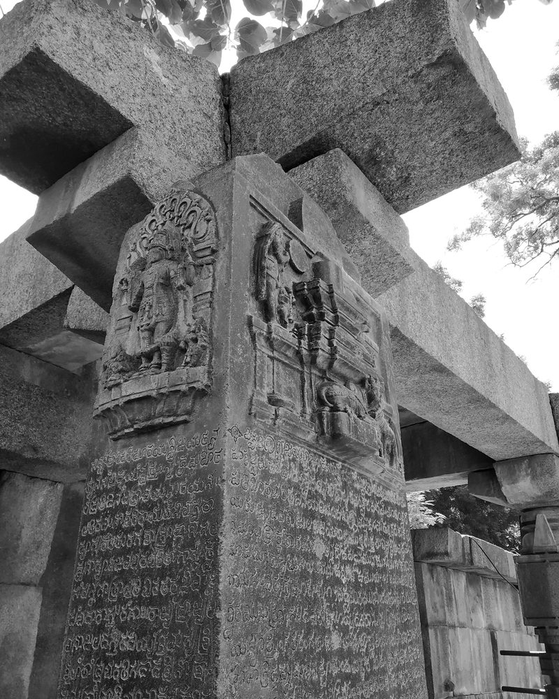 A dark stone pillar under heavy granite beams carries carved panels of gods above many rows of engraved letters.