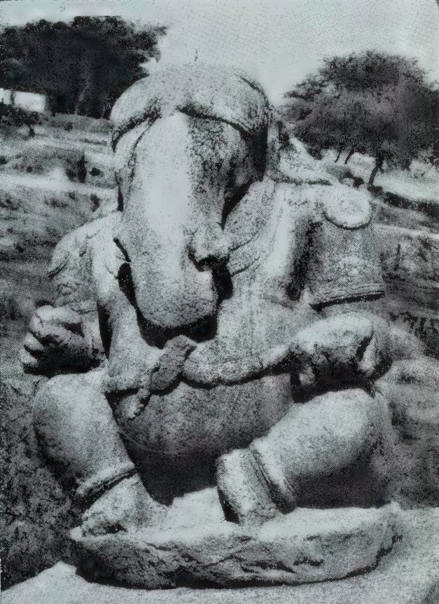 A large seated stone Ganesa with an elephant head and a broad belly, in the open with trees behind.