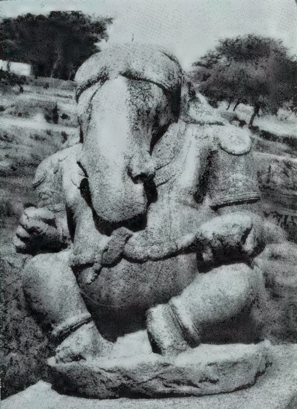 A large seated stone Ganesha with an elephant head and a broad belly, in the open with trees behind.