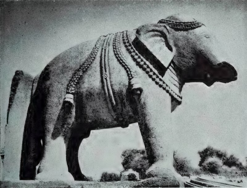 A large stone elephant wearing carved chains, bells and tassels, photographed from a low angle against the sky.