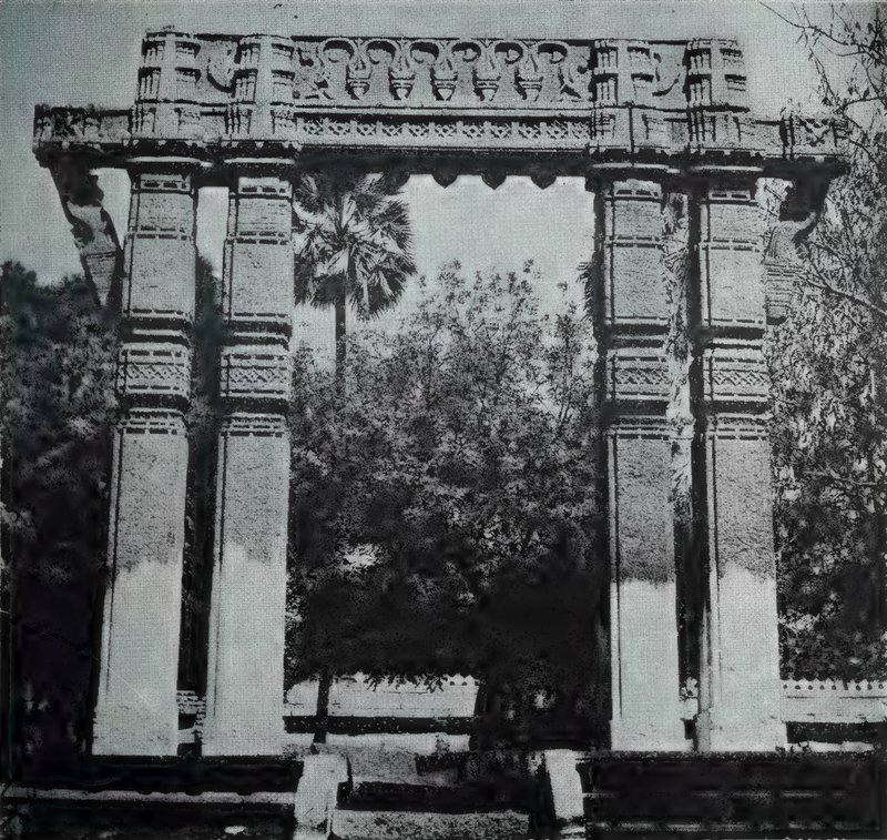 A stone gateway of four tall pillars carrying a carved lintel, with trees and a palm behind it.