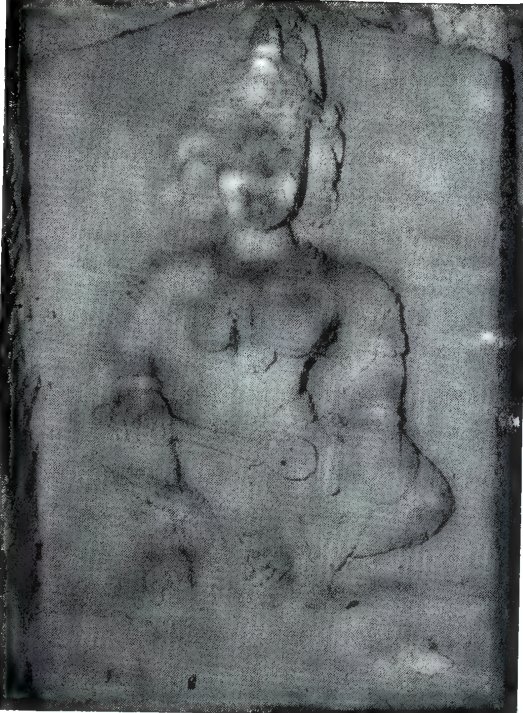 A dark photograph of a seated goddess with a tall crown, carved in low relief on a stone slab with a rounded top.