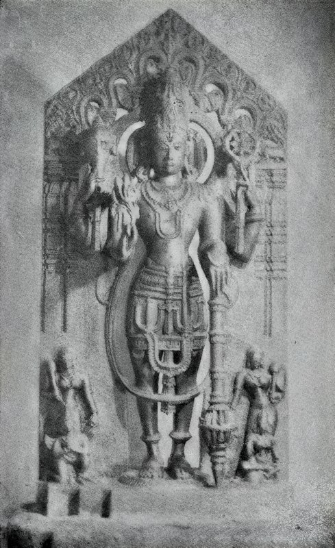 A standing four-armed deity on a stele with a pointed top, with small attendant statues at its feet.