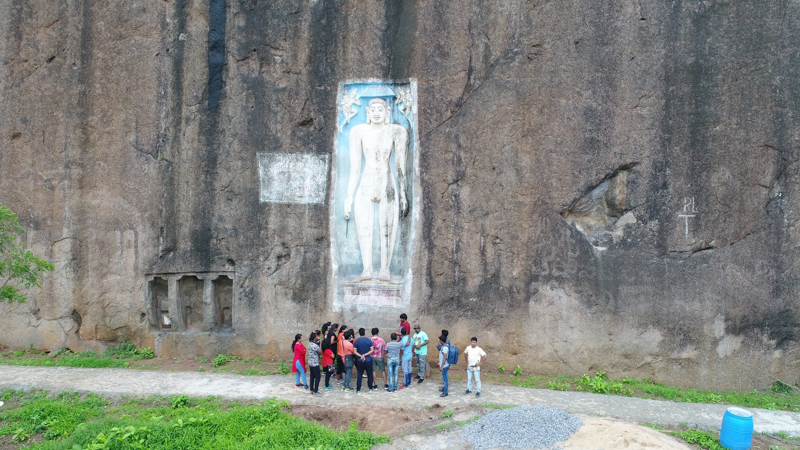 A sheer dark granite rock face with a tall standing Jain sculpture carved in a sunken panel. The statue is painted white on a blue ground. A group of about fifteen visitors stands on a path at its foot. A small opening with square pillars is cut into the rock at the lower left.
