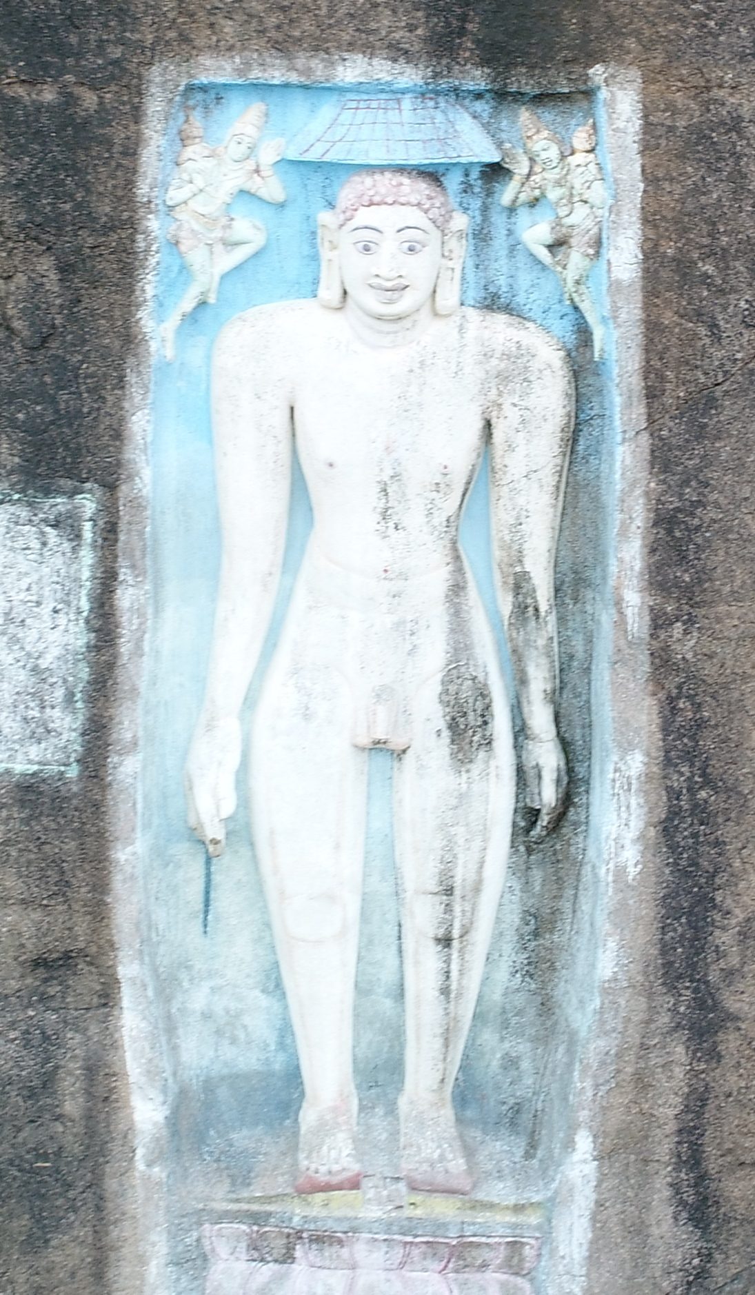 A nude standing Jain sculpture in relief, with the arms hanging straight at the sides. The statue is painted white, with dark curled hair and painted eyes. A single parasol is carved above the head, and a small flying attendant with joined hands is carved in each upper corner.