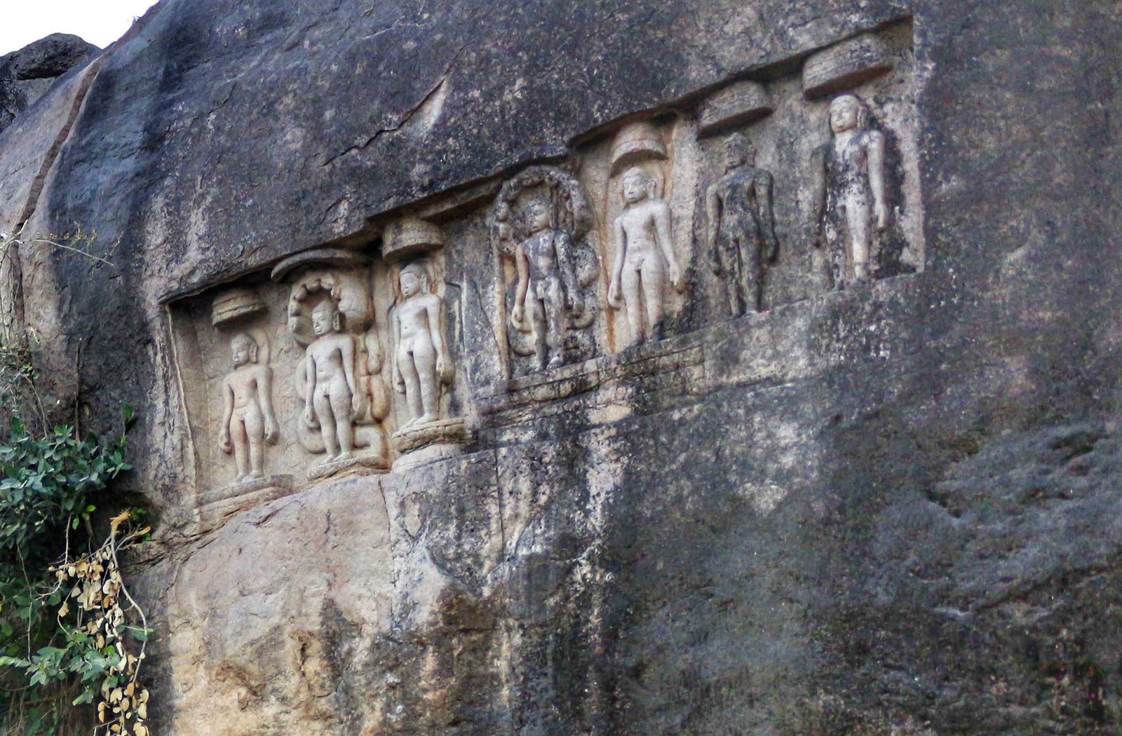 A row of seven small standing Jain sculptures carved in shallow niches along the face of a large grey boulder. Two of the sculptures have the coils and hood of a snake behind them. Parasol shapes are carved above several of the heads.