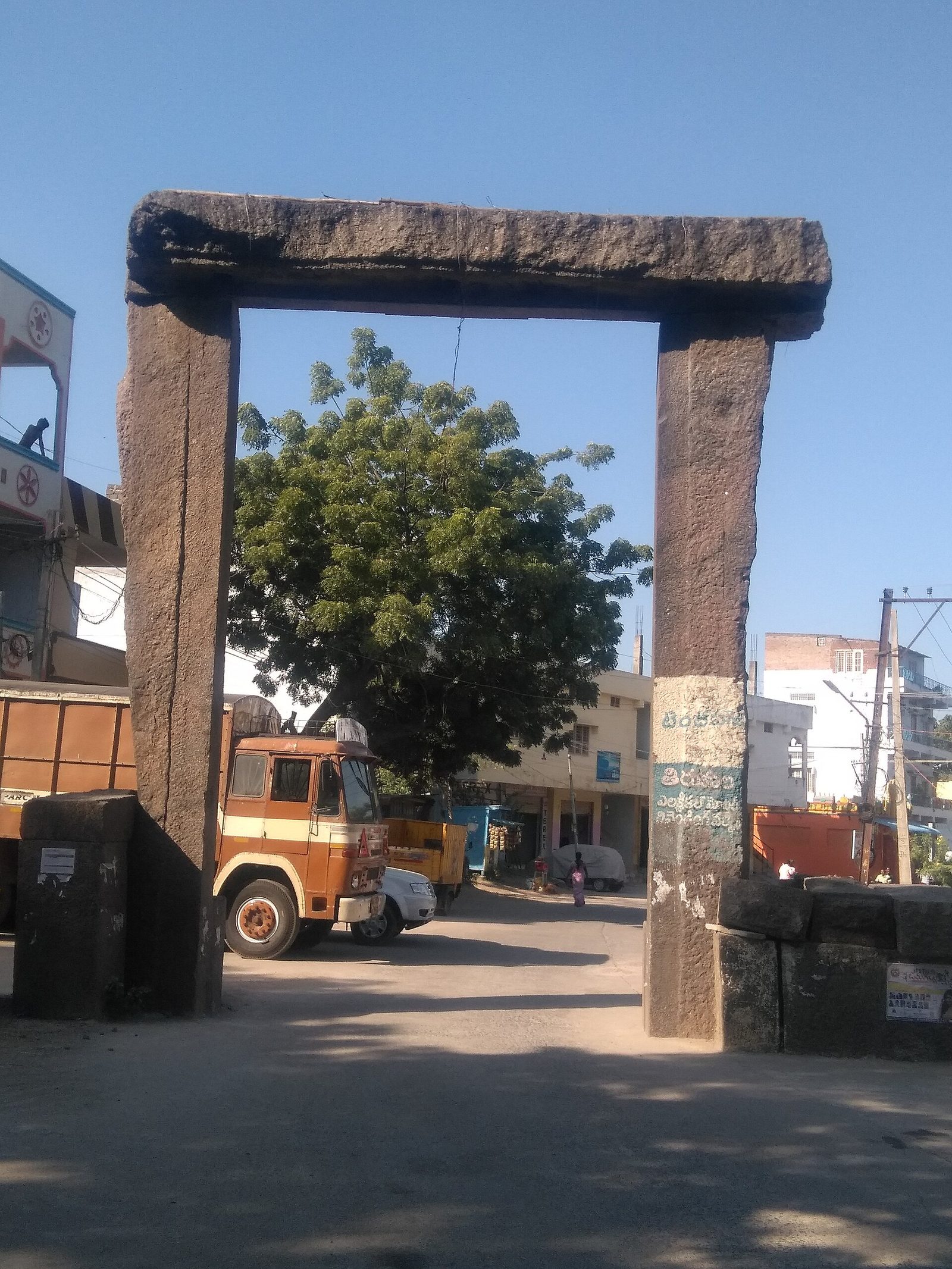 A tall stone gateway at Hanamakonda. Two rough monolithic posts carry a single heavy stone lintel, and a street with trucks and buildings lies beyond.