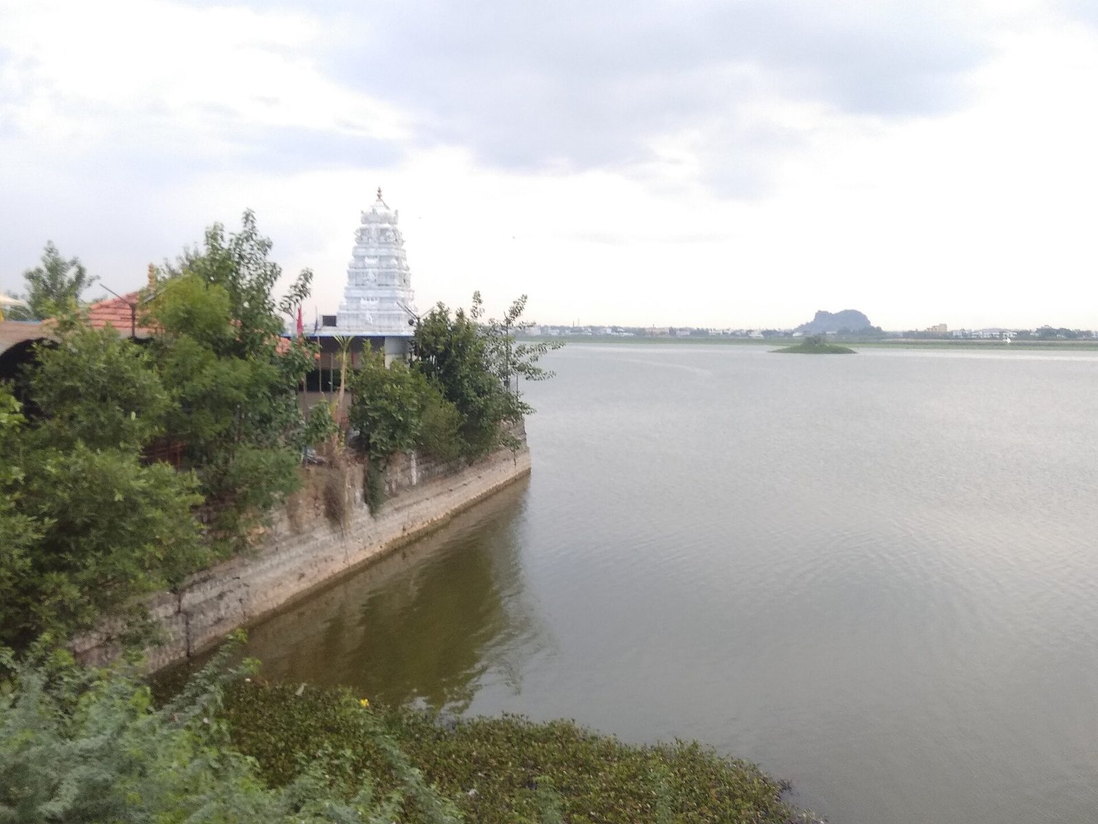 A white tiered temple tower rises above trees on a stone-faced embankment at the edge of a wide lake. A small island and a distant rocky hill are visible across the water under a cloudy sky.
