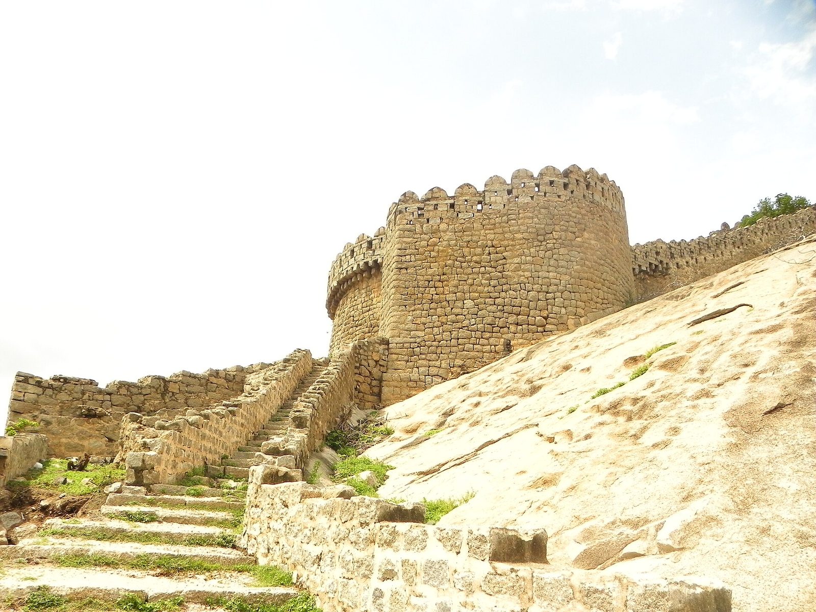 A round stone tower with a notched top edge stands on a slope of bare granite. Stone steps with low side walls climb up beside the tower. A notched stone wall continues along the rock to the right.