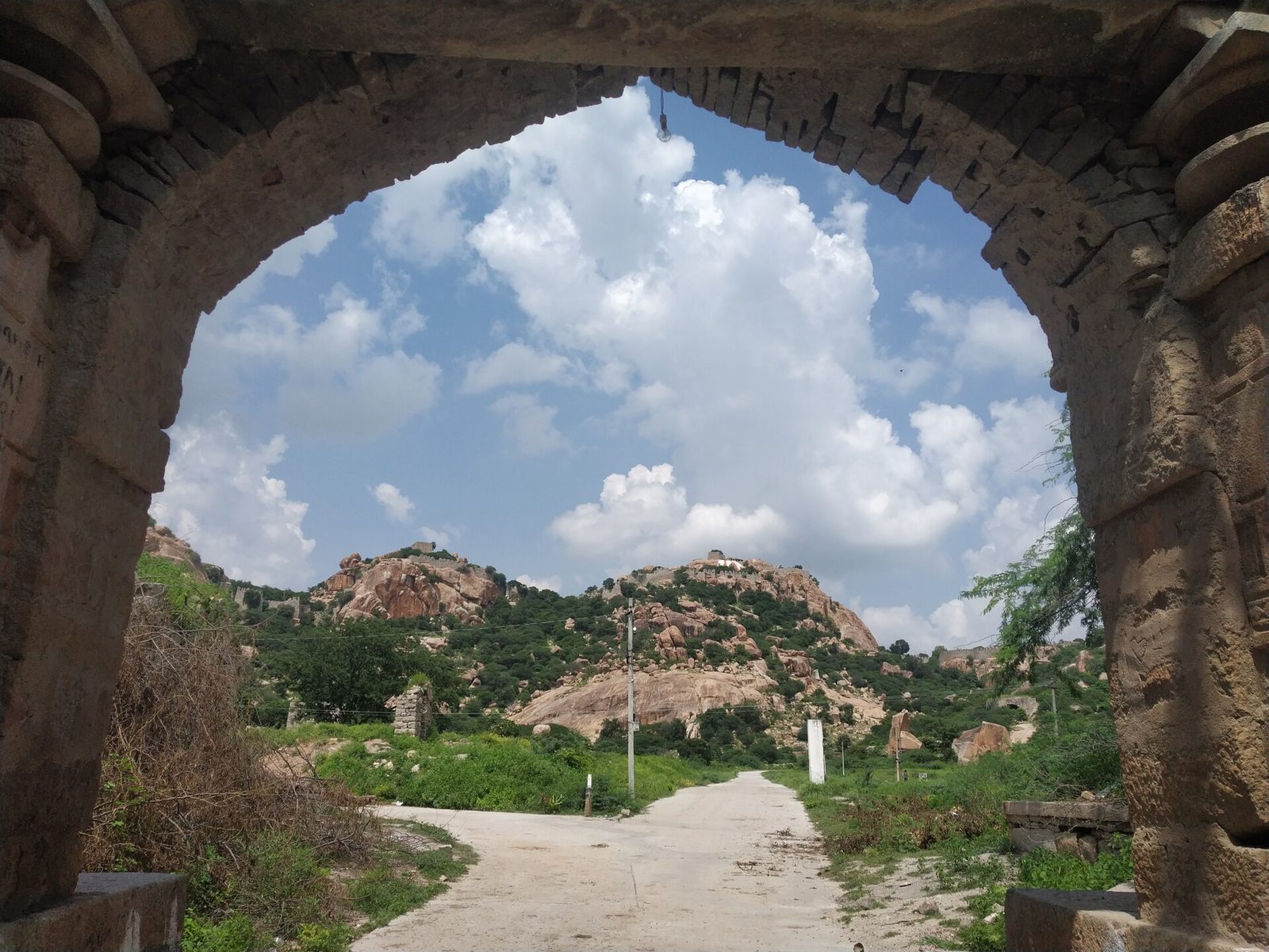 A large stone arch frames a road that leads towards rocky hills under white clouds. Small white structures stand on two of the hilltops. Green scrub covers the lower slopes.