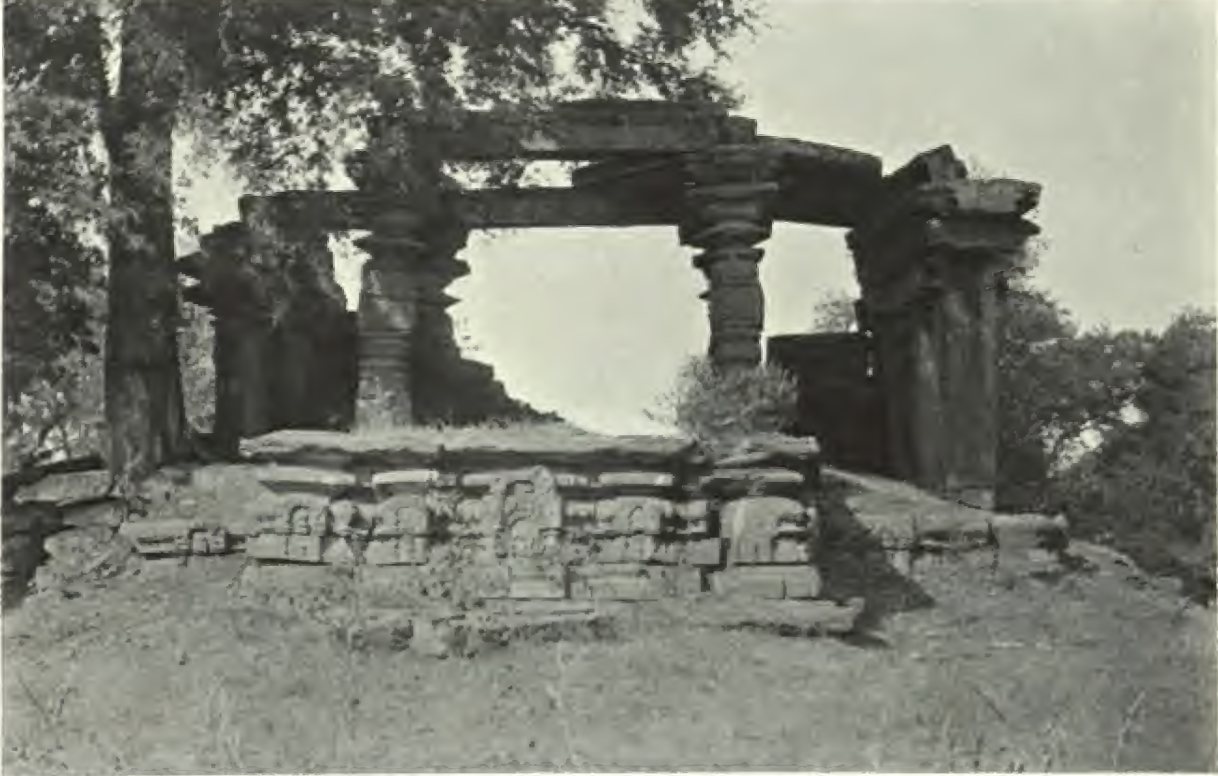A pillared stone porch with turned pillars and heavy beams stands on a carved platform, with trees behind it.