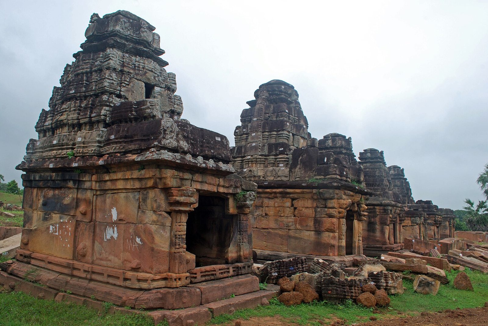 A row of small sandstone shrines at Ghanpur, each a square cell with a stepped tiered superstructure, receding into the distance, with fallen blocks on the grass