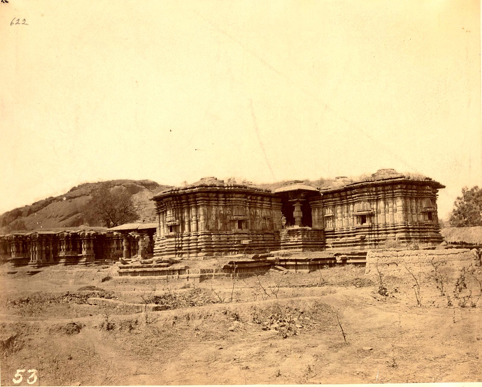 The square stone shrines of the Thousand Pillar Temple stand in front of a rocky hill. A long pillared hall extends to the left. Mounds of earth and scrub lie in front.