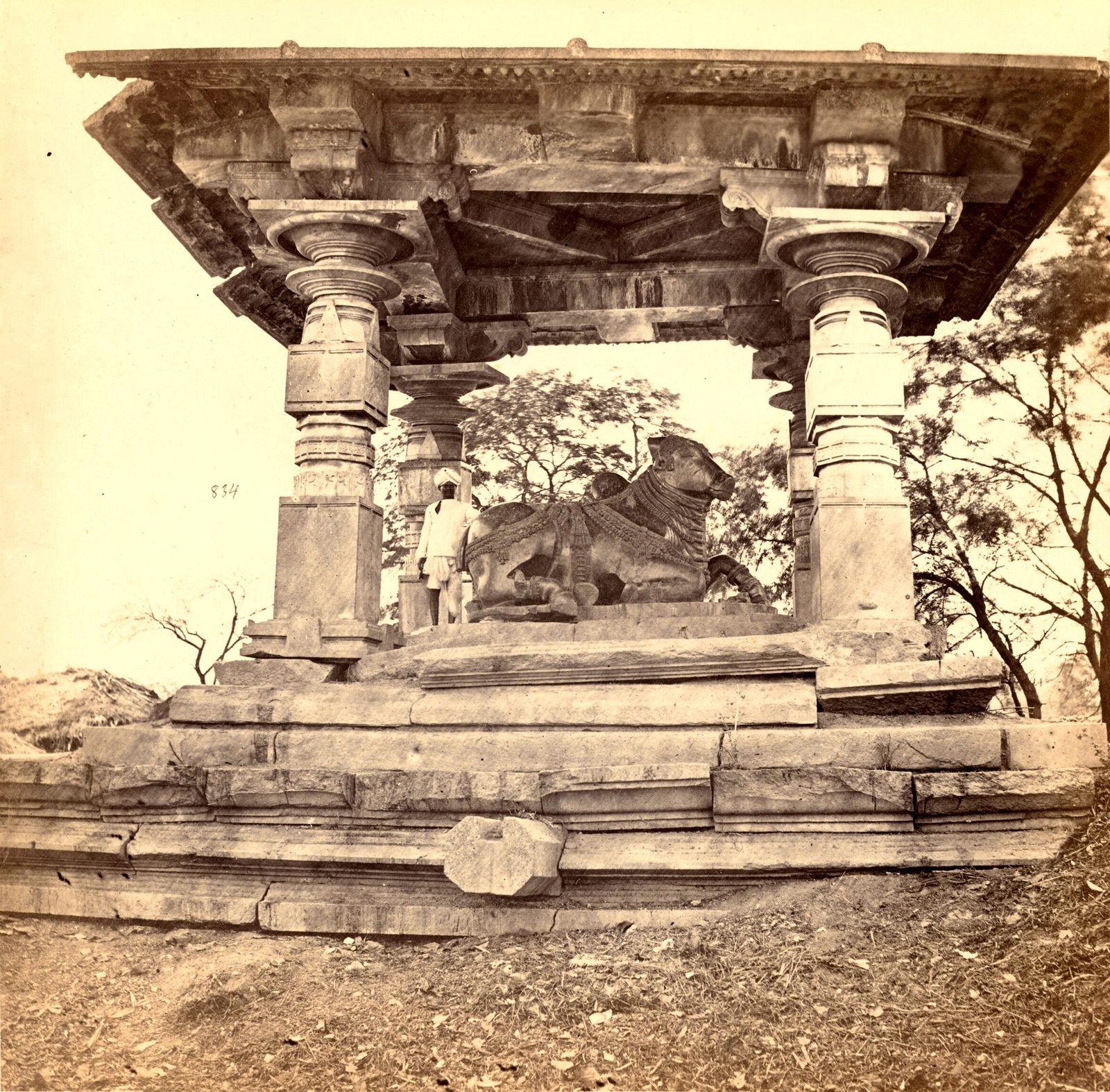A carved stone bull lies under a flat stone roof held up by round carved pillars. The porch stands on a stepped stone platform. A man stands beside the bull.