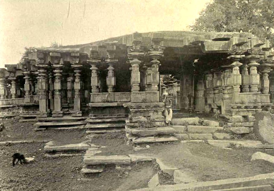 A large hall with many carved stone pillars under a flat roof. The roof line sags, and some roof stones are missing. Displaced stones and slabs lie in front of the hall.