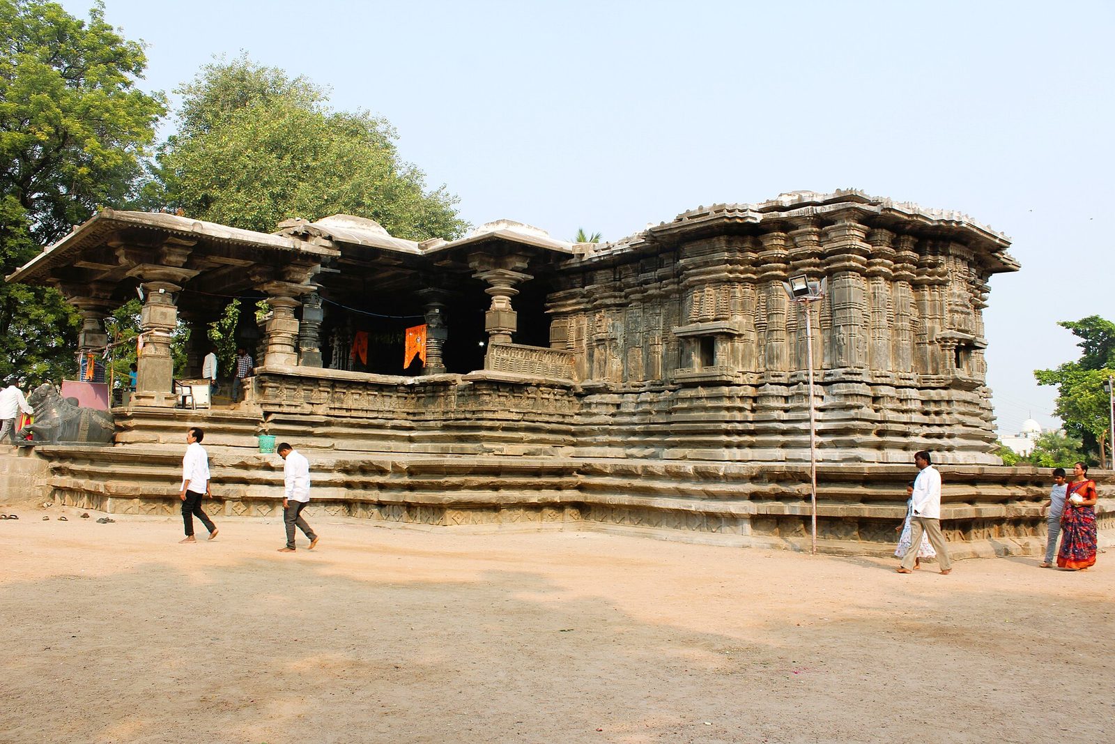 Exterior of the Thousand Pillar Temple at Hanamakonda: a dark stone shrine block with densely moulded walls at right and an open pillared porch at left, all on a broad stepped platform