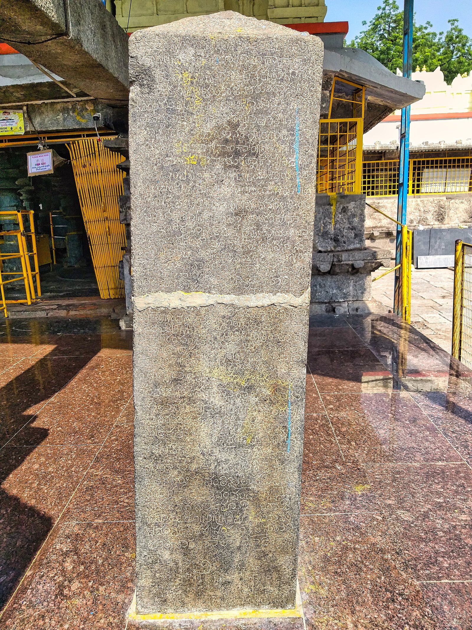 A tall square stone pillar is covered with worn lines of Telugu letters. A crack across the middle has been filled with mortar.