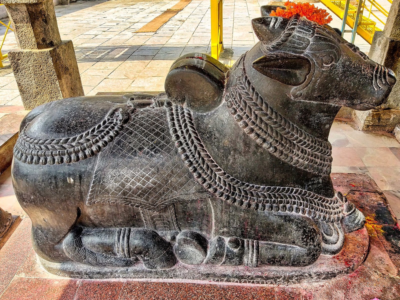 A seated bull carved in dark polished stone wears chains of bells and a cloth with a cross-hatched pattern. Flowers lie on its head.