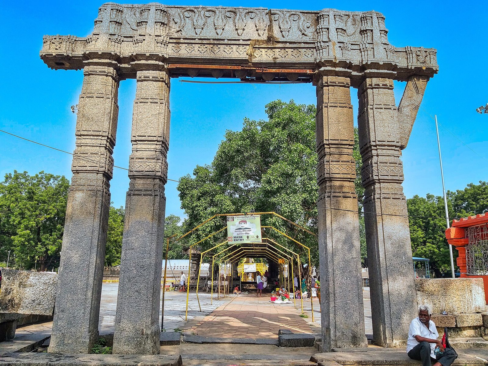 A tall stone gateway of two pairs of square pillars carries a broad carved beam. A paved path runs through it towards the temple.