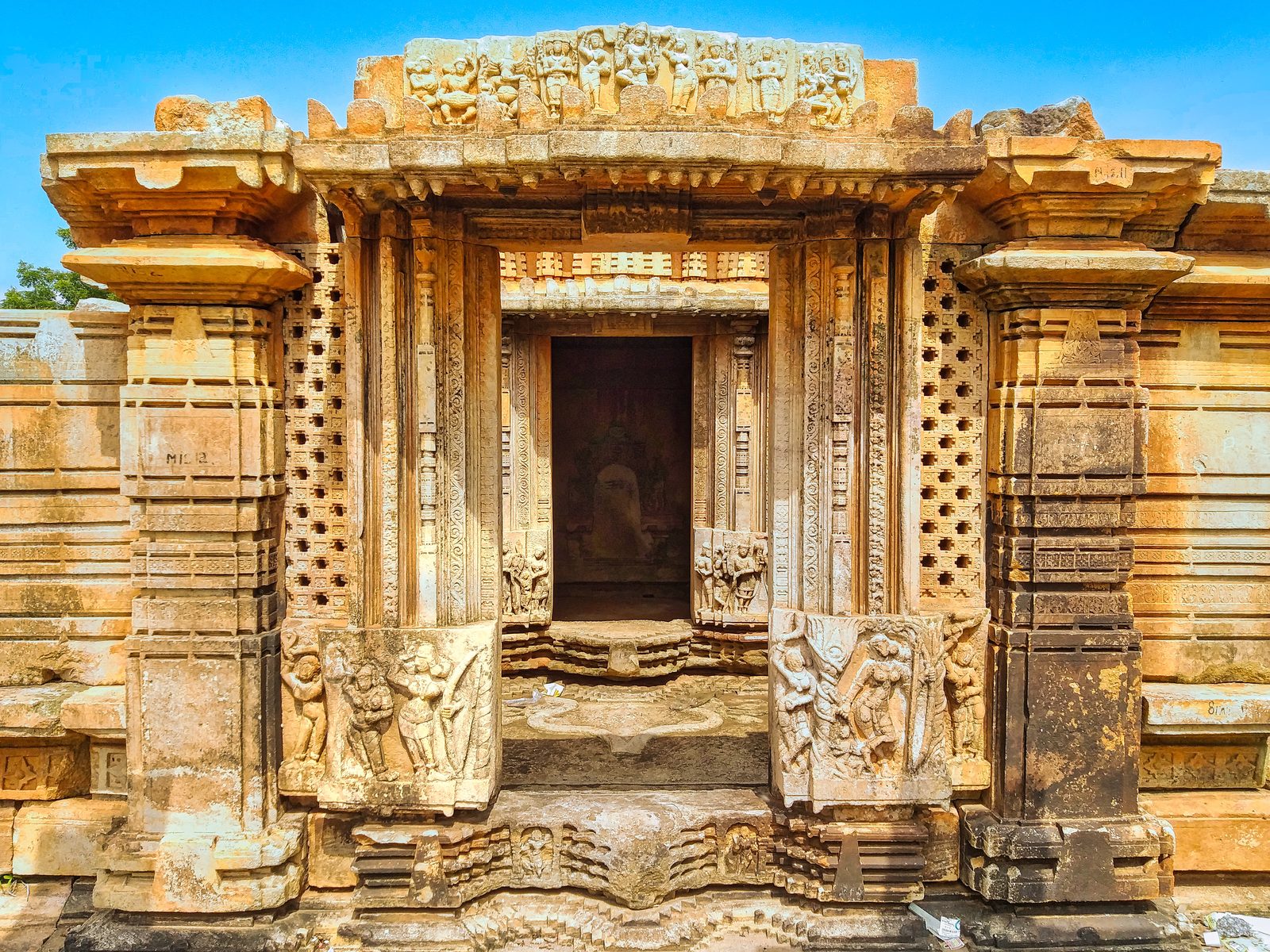 A carved stone doorway leads into a dark inner chamber. Bands of carved scrolls and small sculptures run up the door frame. Perforated stone screens stand on both sides of the door. Panels with standing people in relief sit at the foot of the frame. A row of small seated sculptures is carved above the door.