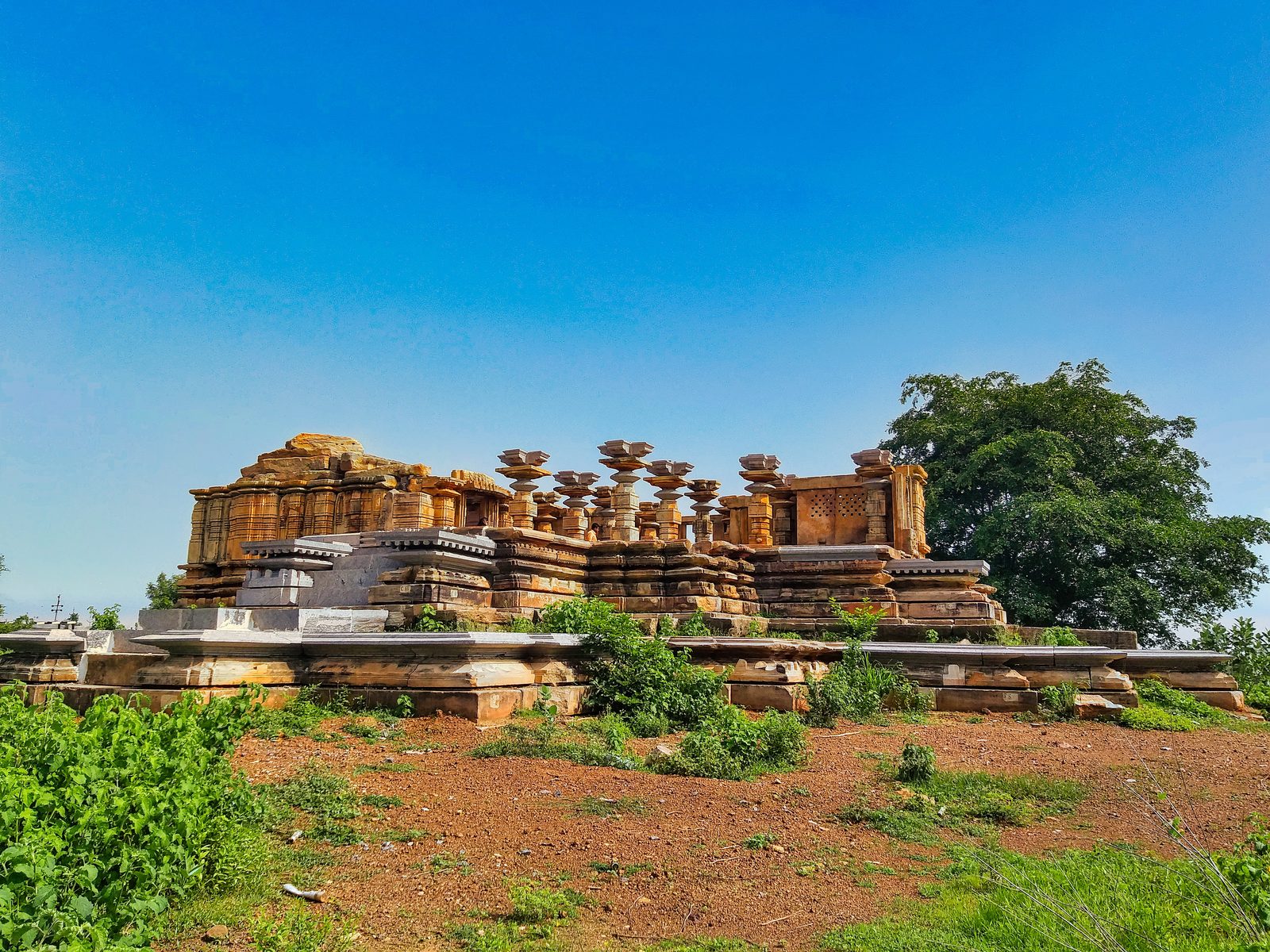 A stone temple stands on a raised platform in open red ground under a blue sky. The shrine walls are partly rebuilt. A row of pillars without a roof stands in front of the shrine. A large tree grows on the right side.