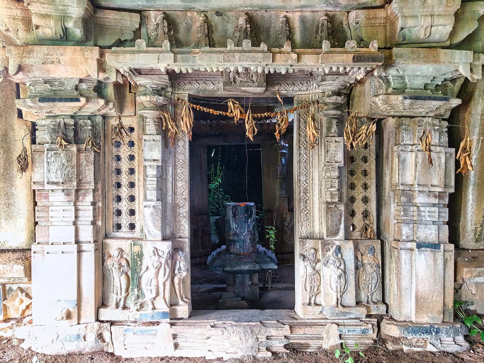 A carved stone shrine doorway between two pierced stone screens with small square openings. Small carved sculptures stand at the base of each door frame. A stone linga is visible in the dark shrine beyond.