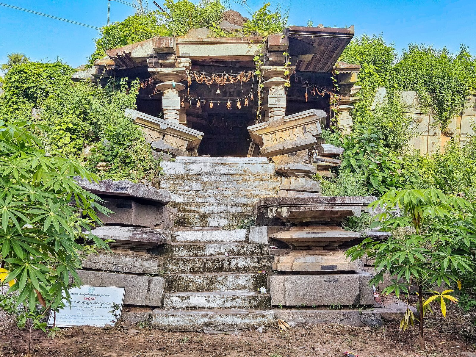 A ruined granite temple on a high base, overgrown with plants. A flight of stone steps leads up to a pillared porch. Displaced roof slabs and beams lie tilted on each side of the steps.
