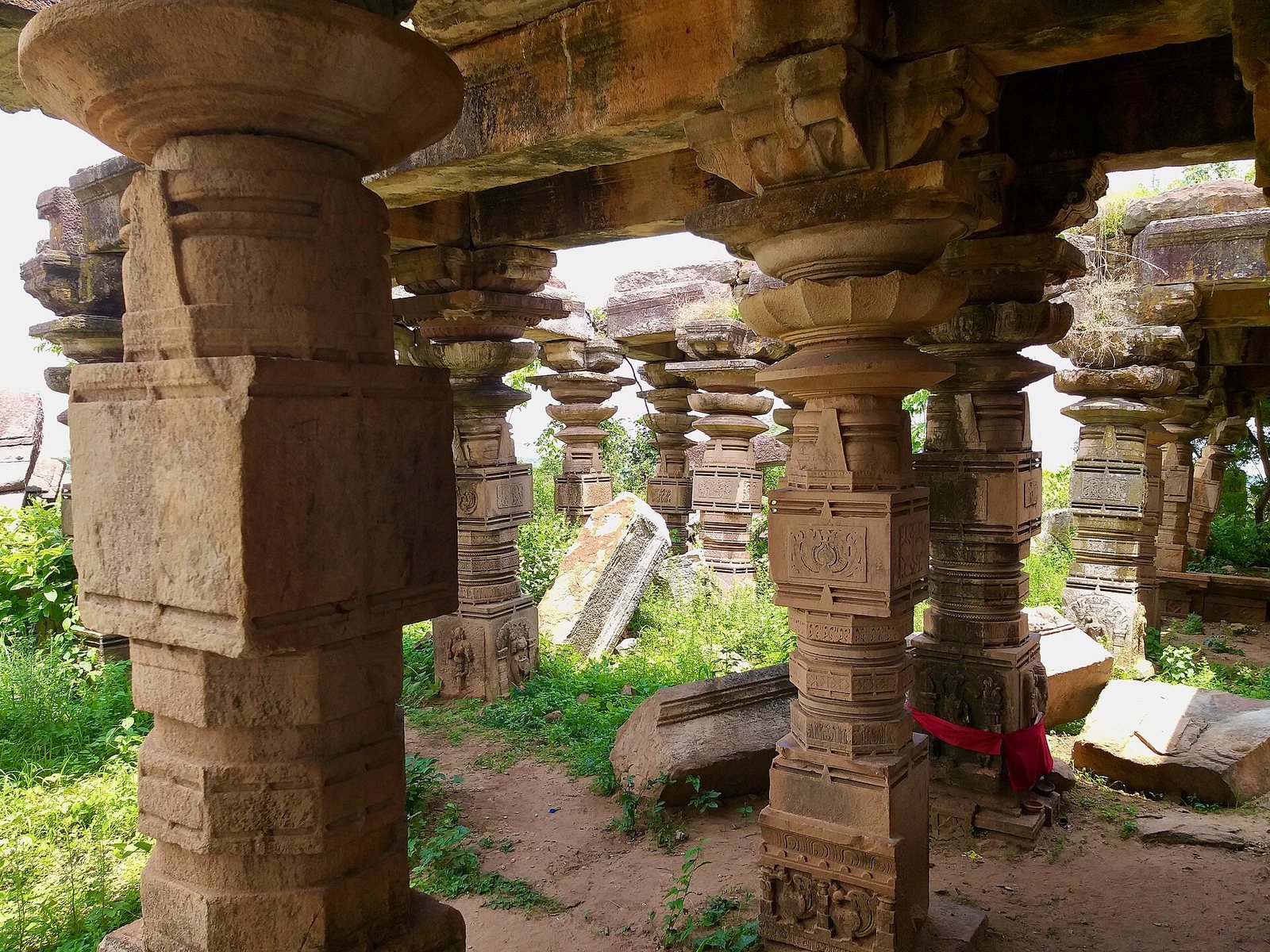 Rows of heavy stone pillars with turned and carved sections hold up flat stone beams. Daylight comes in between them, and broken stones lie on the floor.