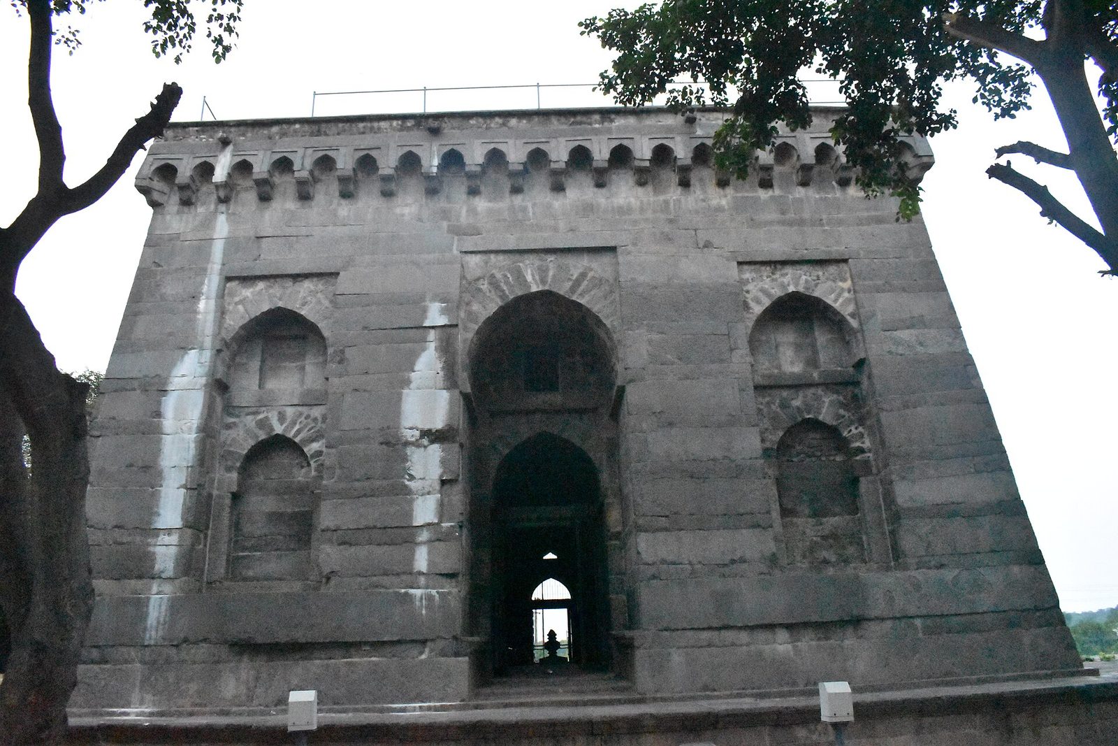 The end wall of Khush Mahal at Warangal: a massive grey stone facade with strongly sloping sides, a tall pointed-arched entrance and arched niches, crowned by a row of small merlons