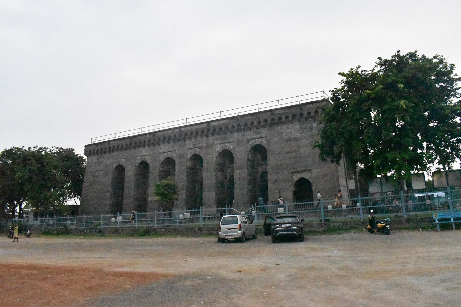 The long side of Khush Mahal, a large rectangular stone hall with a row of tall arched recesses, seen across open ground with parked cars