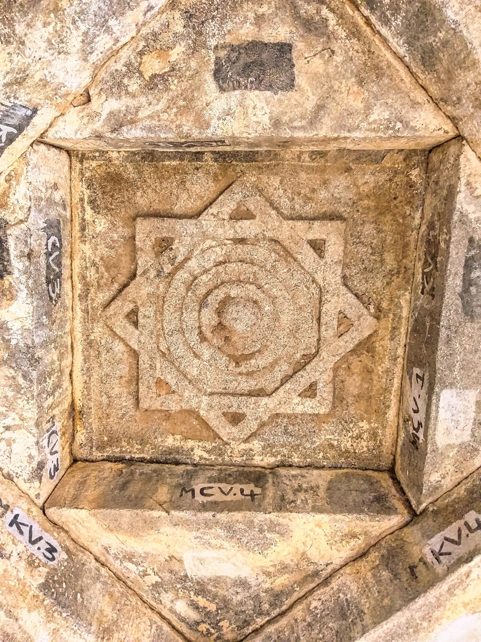 A square stone ceiling panel carved with an eight-pointed star around rings, framed by sloping slabs with painted numbers.
