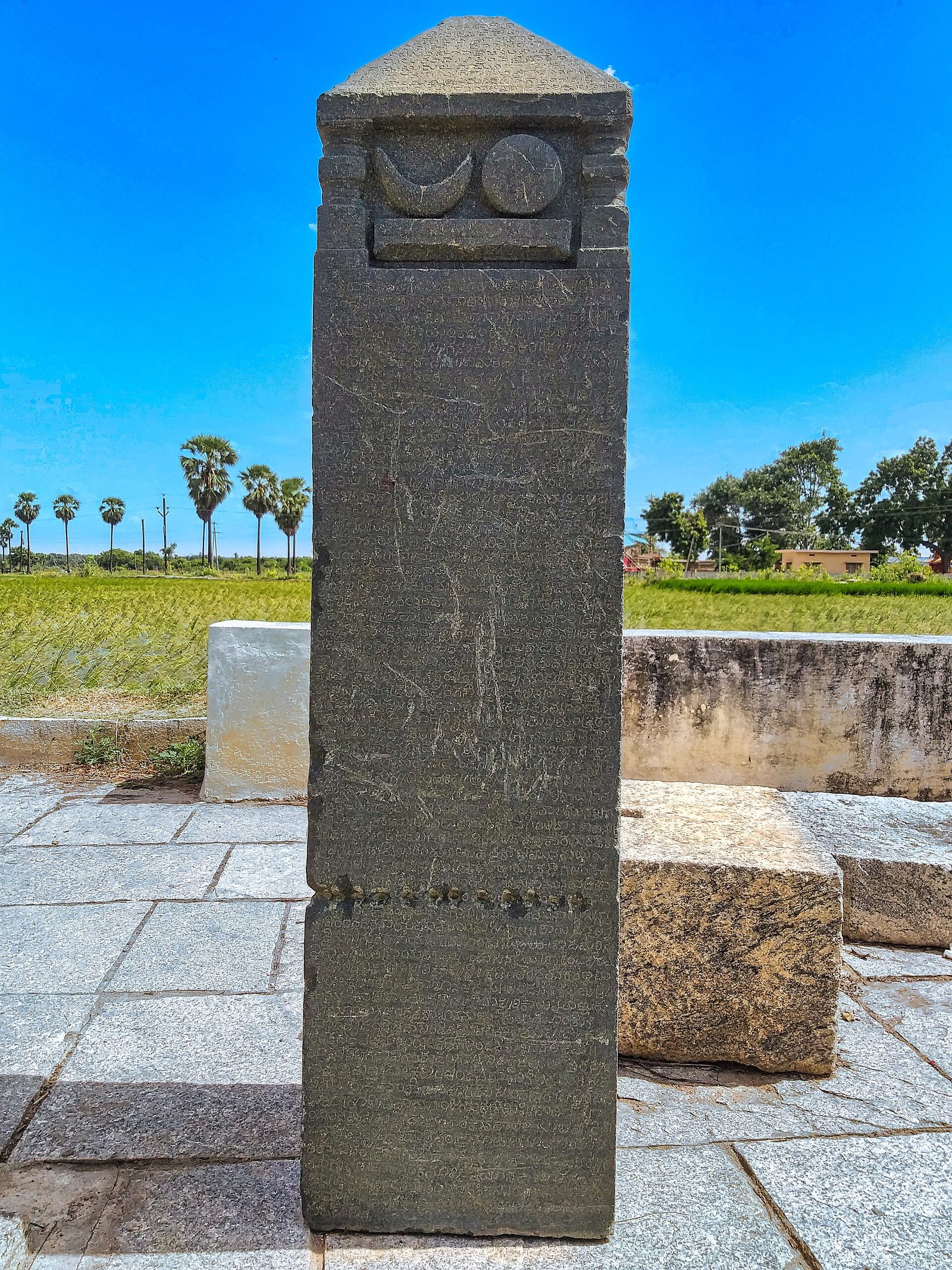 A square stone pillar inscribed on its faces, topped by a carved crescent moon and sun and a pyramidal cap, standing on a paved platform