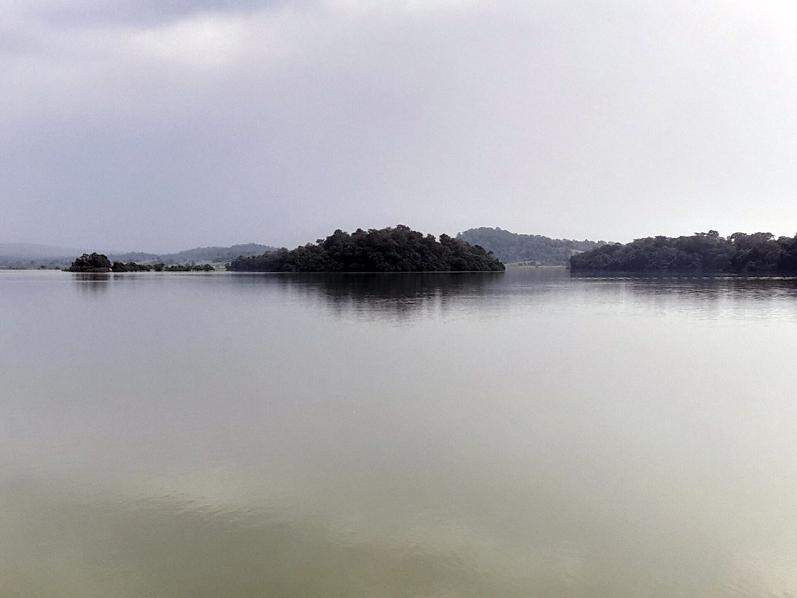A broad, still expanse of lake water under a grey, overcast sky. Several low, tree-covered islands lie across the middle distance, with faint hills on the horizon.