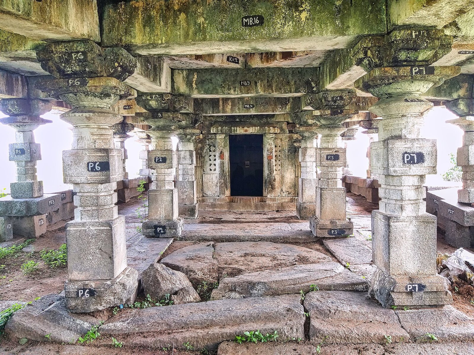 Rows of square stone pillars stand in a hall with a flat stone ceiling. At the far end, a dark doorway between two pierced stone screens leads into the sanctum.