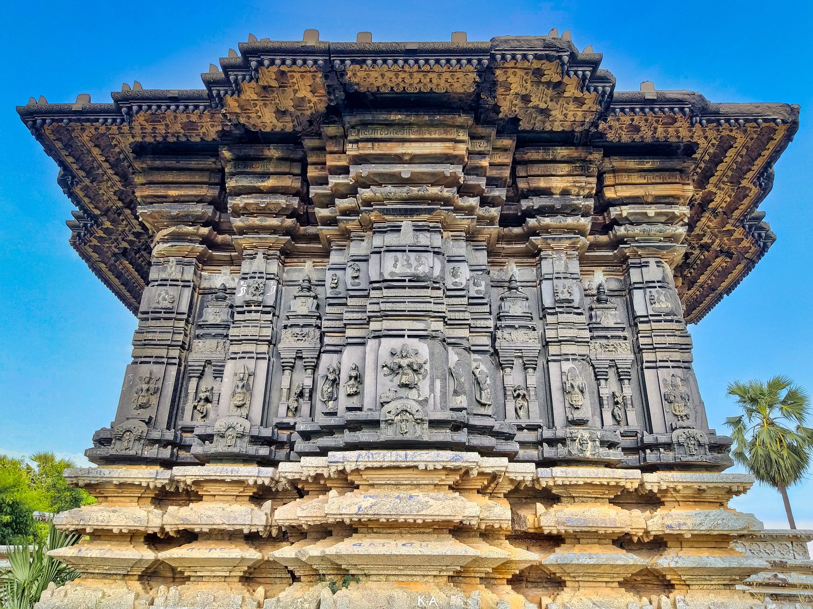 A dark stone wall is divided by many projecting pilasters, each carved with small gods in niches. A broad overhanging cornice runs along the top, above a pale stone base of mouldings.