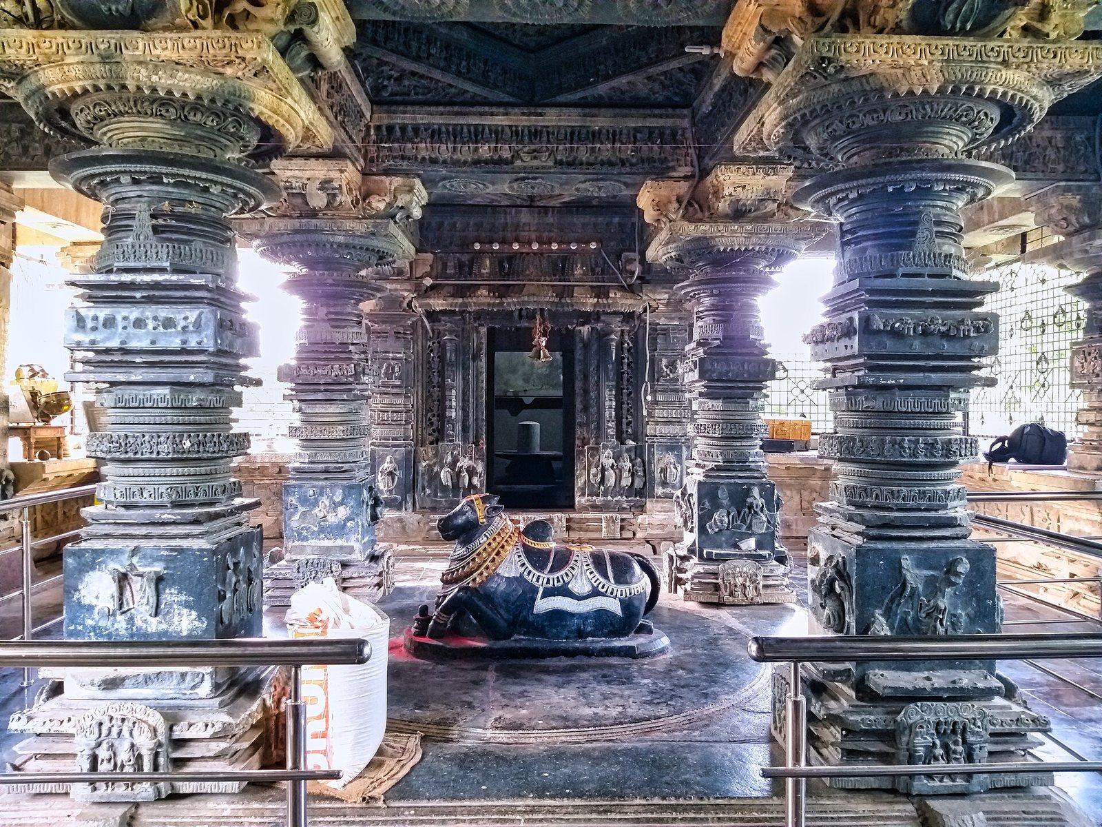 Inside a temple hall, heavy dark stone pillars with turned and carved bands stand around a seated stone bull. A shrine doorway is visible behind, and metal railings surround the pillars.