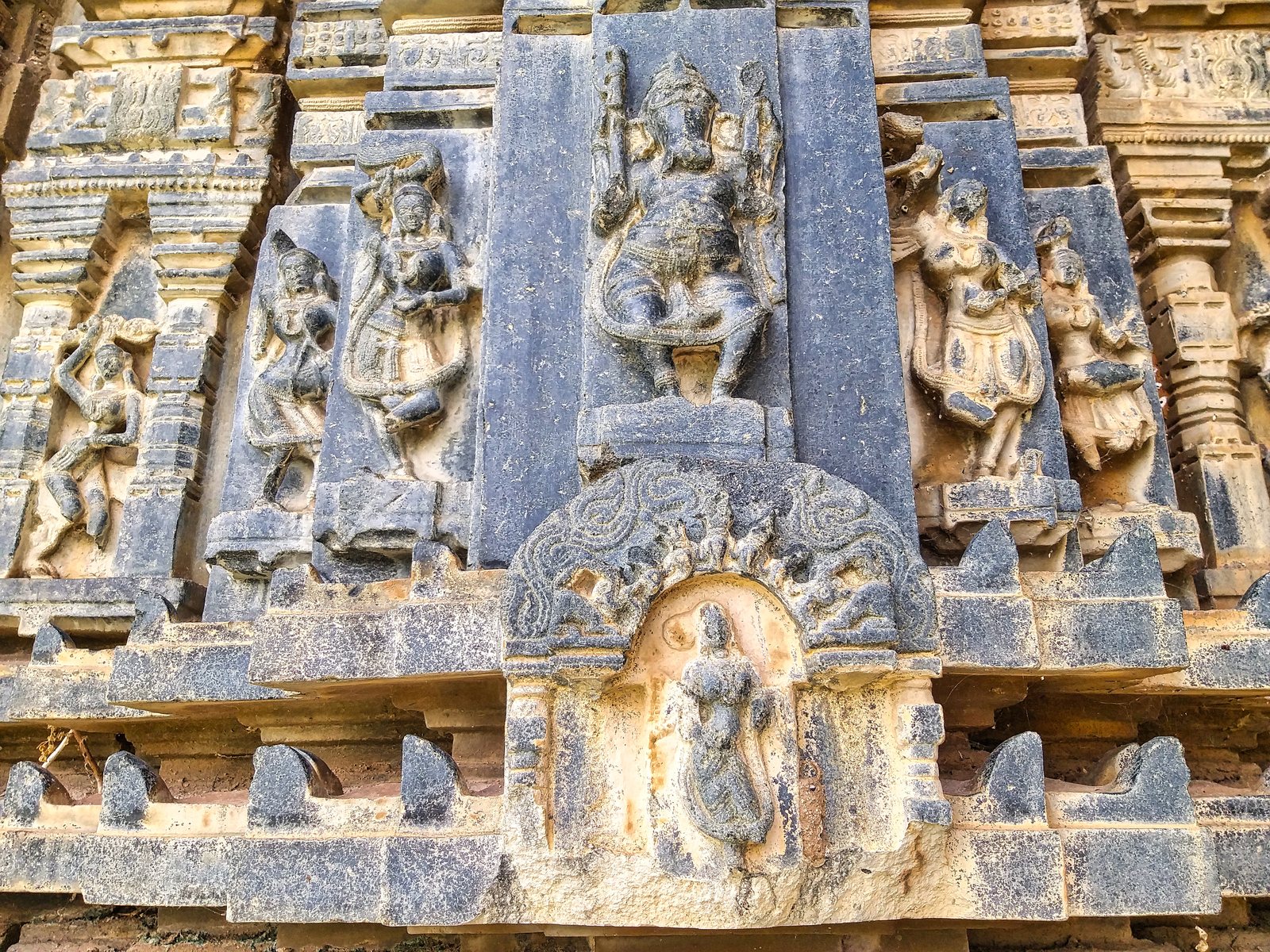 Close view of weathered carving on a temple wall. An elephant-headed god stands in the centre with dancers on either side. Below him a small woman stands inside a carved arch.