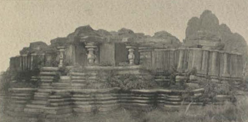 A ruined temple stands on a broad platform of many stepped stone layers. Turned pillars stand at the front, and a rocky hill rises behind on the right.