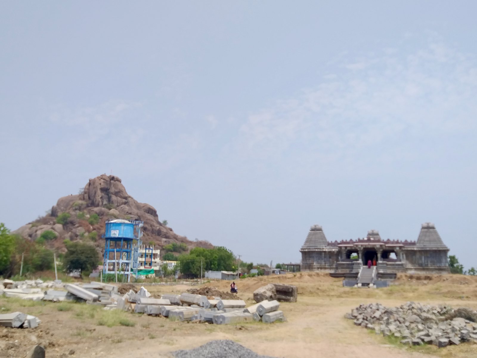 A grey stone temple with three towers stands on a platform with a long flight of steps. A rocky hill and a water tank rise on the left, and cut stones lie in the foreground.
