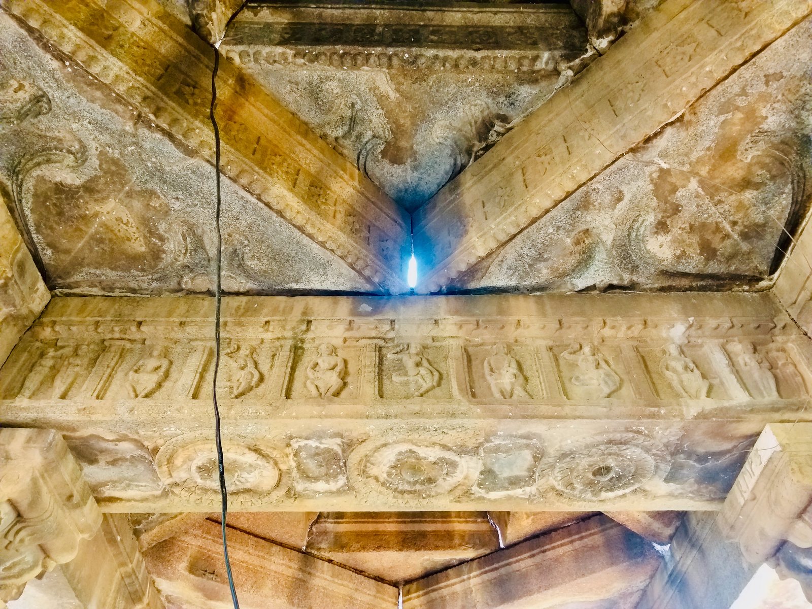 A stone beam seen from below. Small panels along its face hold carved dancers and musicians, and three large lotus flowers are carved on its underside.
