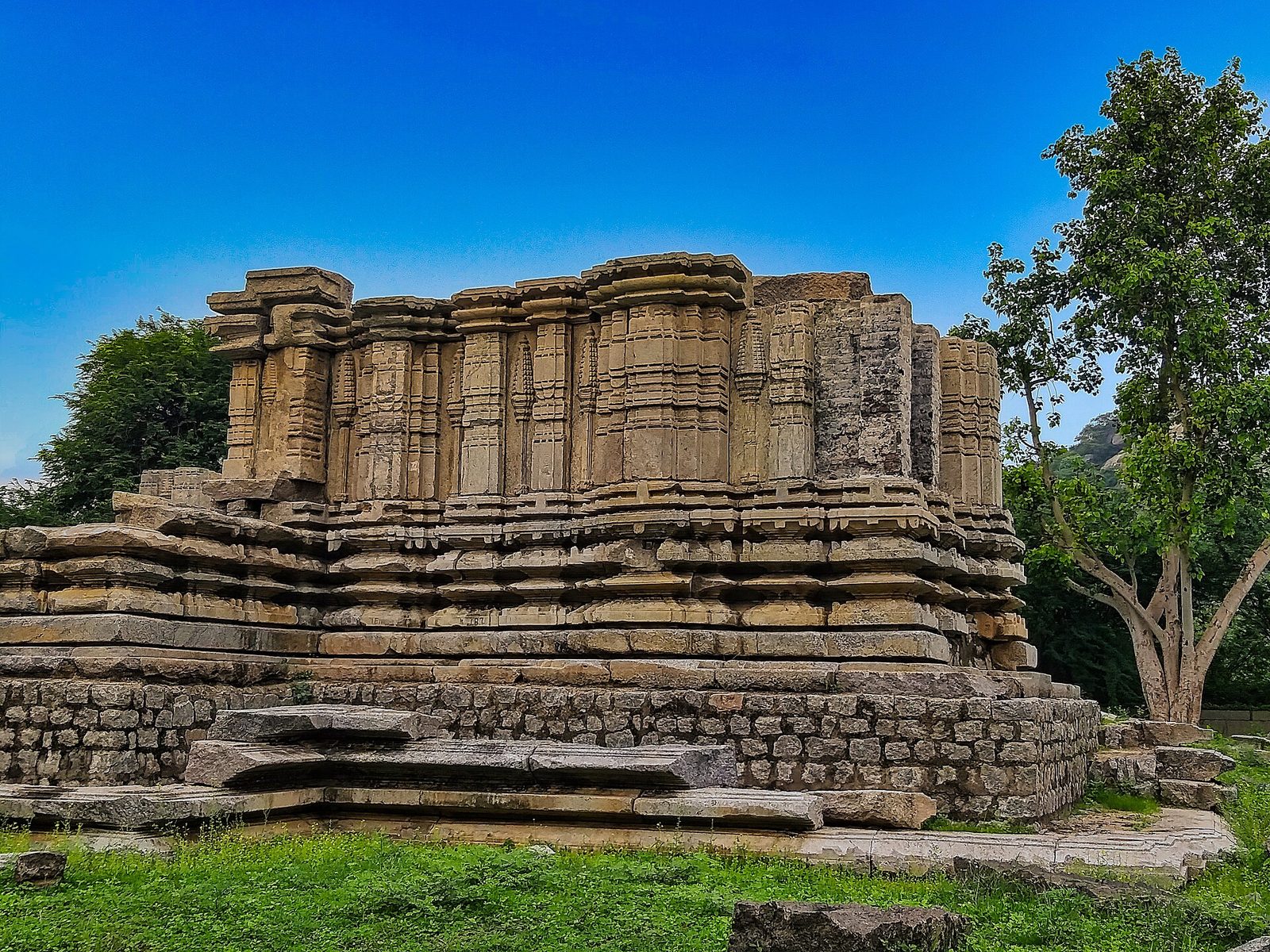 Ruined walls of a stone temple at Nidigonda standing on a moulded base, the superstructure lost, beside a tree under a blue sky