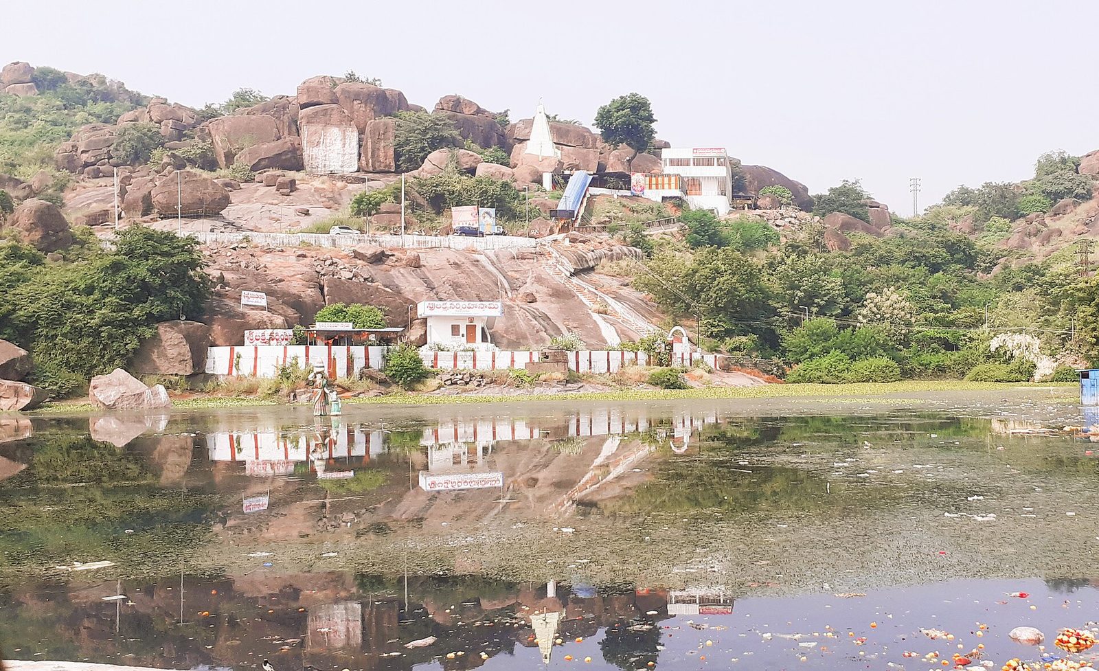 A rocky granite hill under a pale sky. A small white pyramidal temple tower stands near the top, with white walls and small buildings on the slope below. A pond in the foreground reflects the hill.