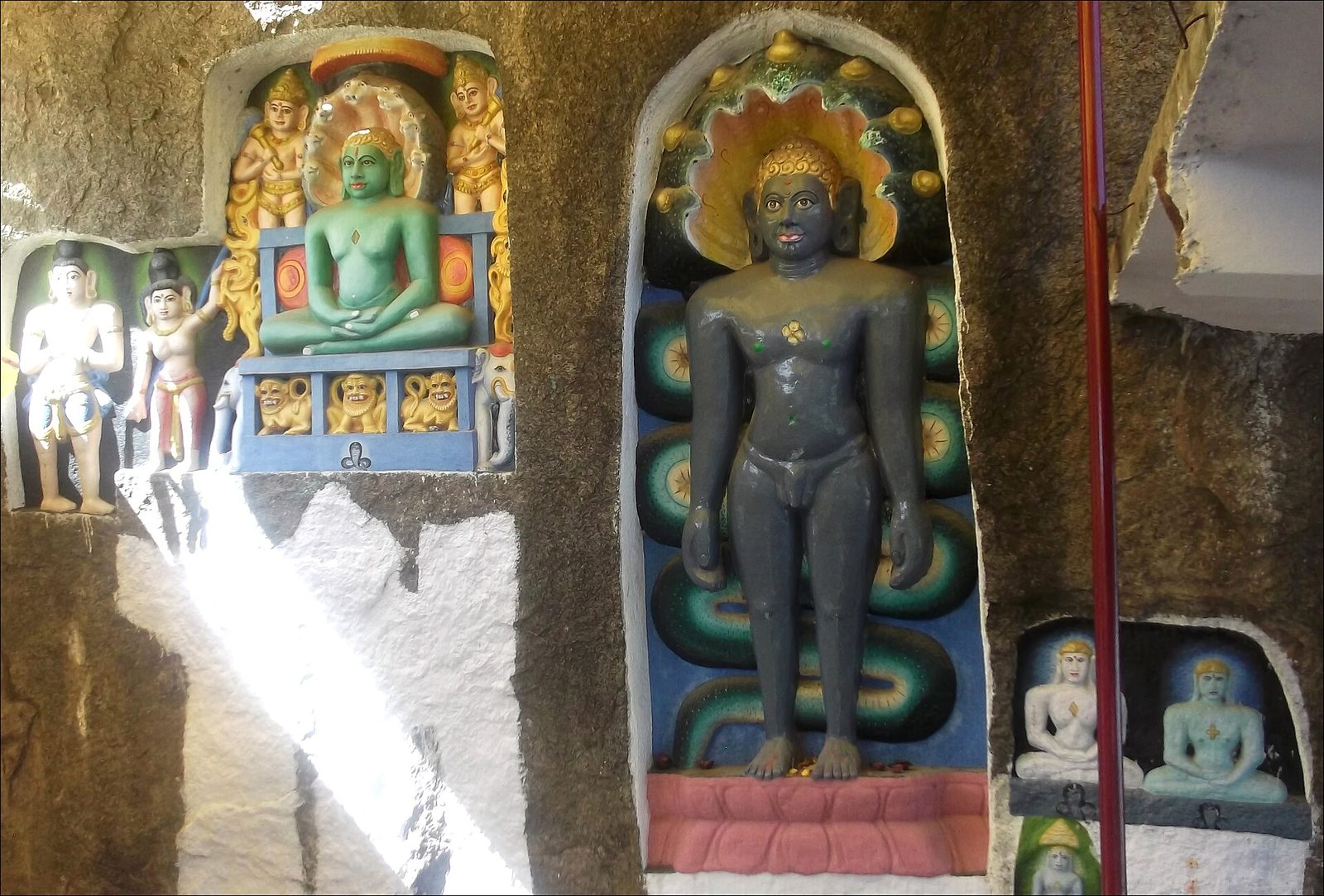 Sculptures carved in niches in a dark rock face and painted in bright colours. A tall standing sculpture painted dark grey stands under a hood of snake heads. A seated sculpture on a throne fills the niche to its left.