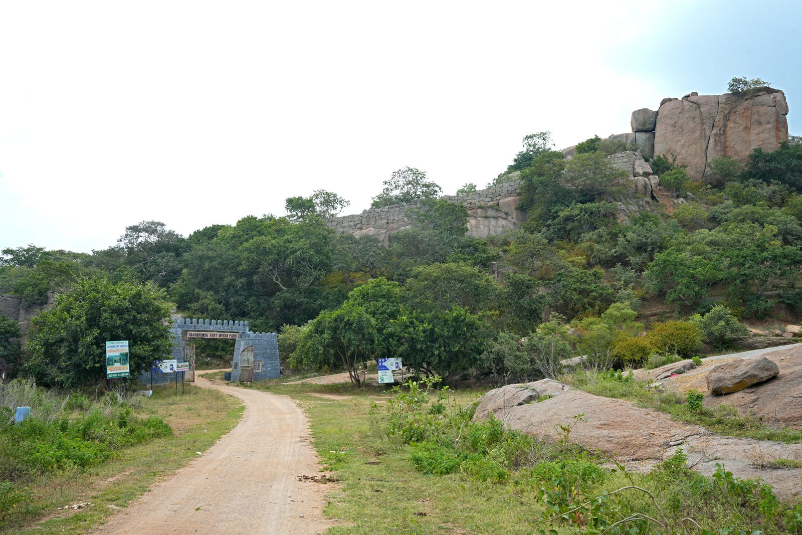 A dirt road leads to a modern stone gate with two square towers at the foot of a wooded hill. Large granite boulders and a stretch of old stone wall stand near the top of the hill.