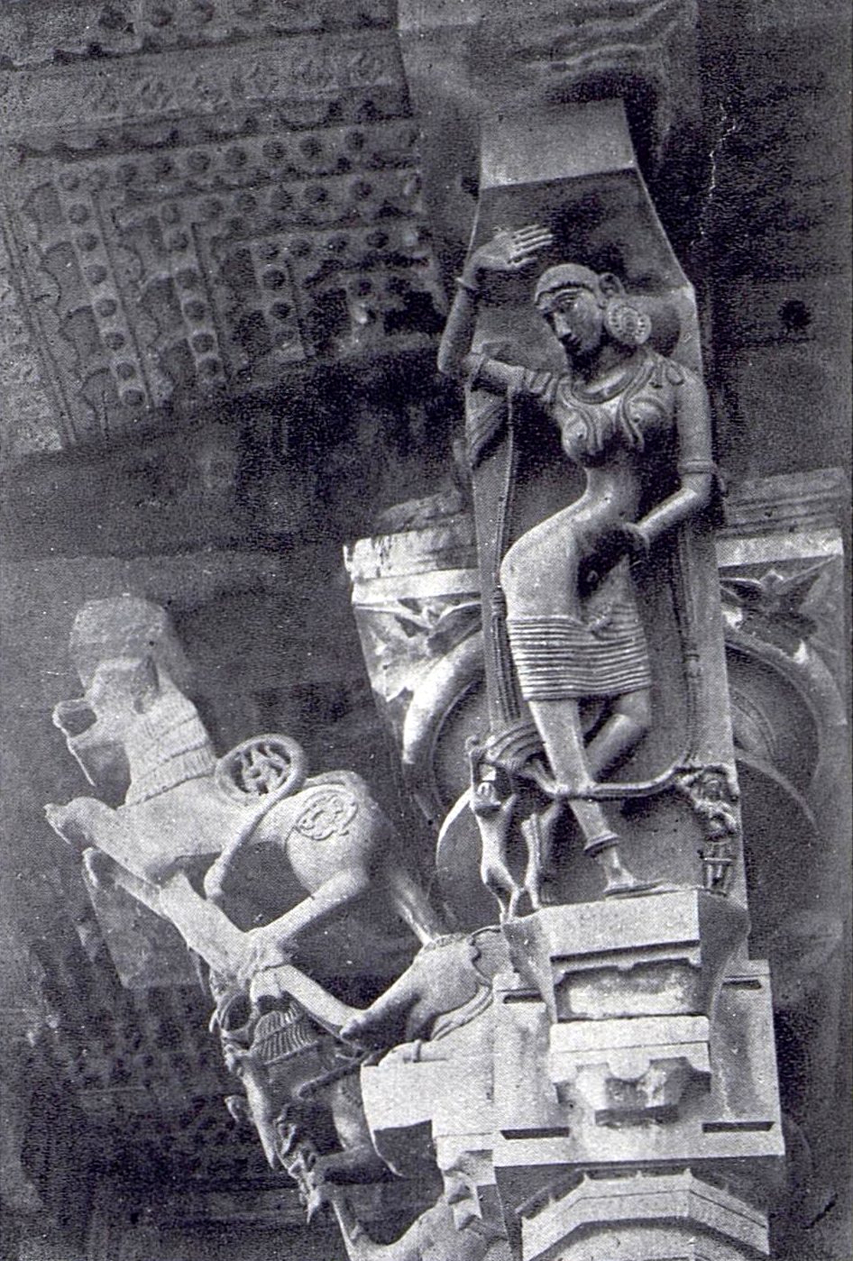A slender stone sculpture of a woman in a dancing pose stands on a sloping bracket under the eave. A sculpture of a rearing beast stands on the next bracket at the left.