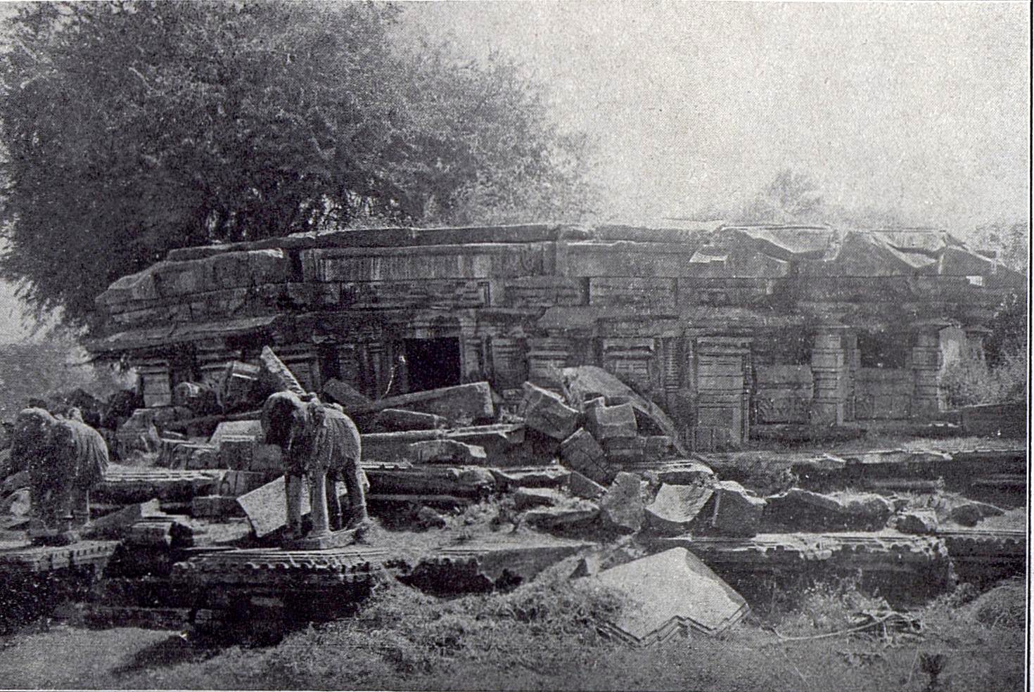 The low walls of a ruined stone temple, with fallen blocks heaped in front of them. Two stone elephant sculptures stand among the fallen blocks. Trees grow behind the temple.