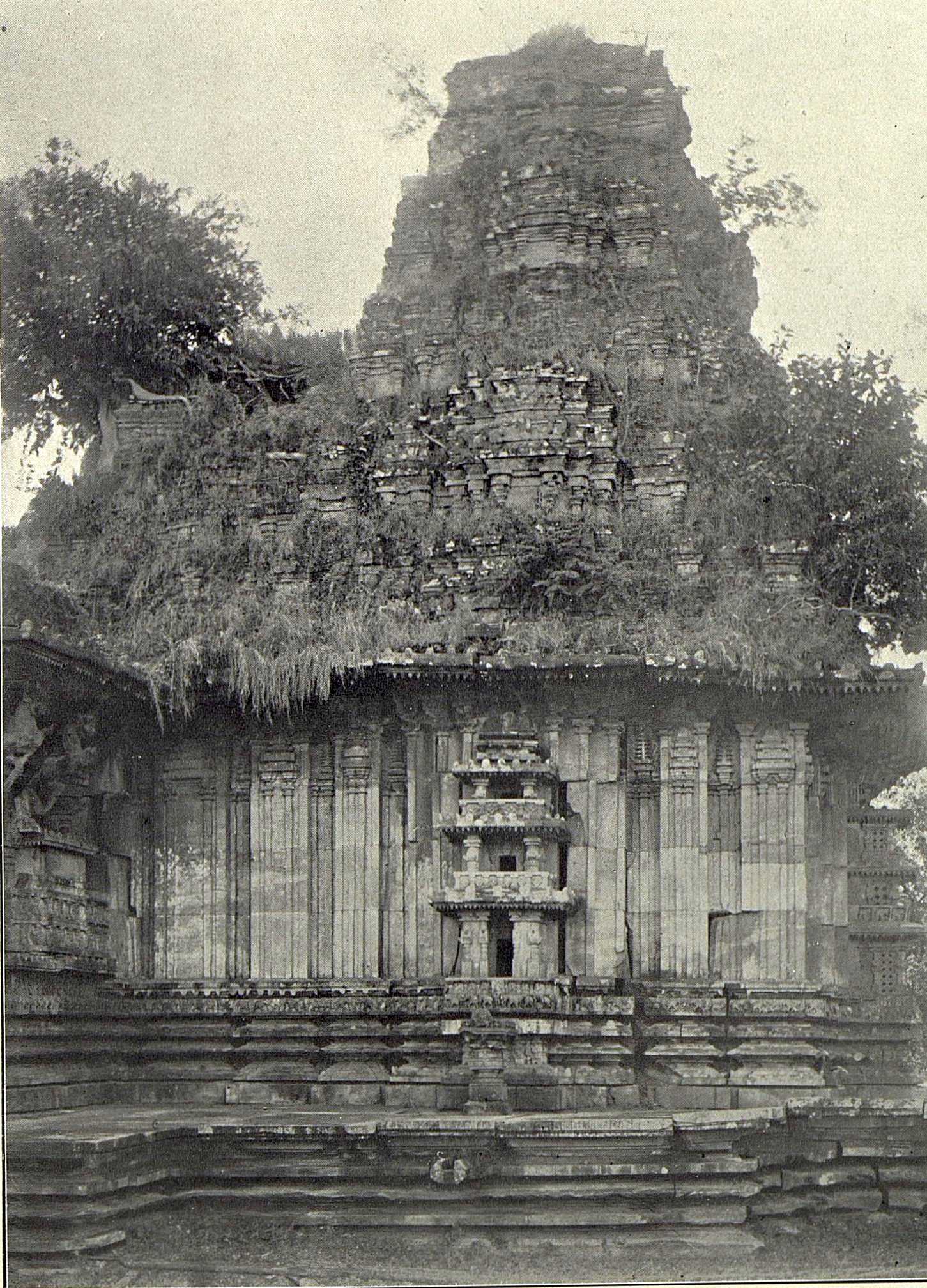 Close view of the stone tower above the Ramappa shrine. Grass and shrubs grow out of the tower, and its upper part is uneven. A small carved niche sits in the shrine wall below.